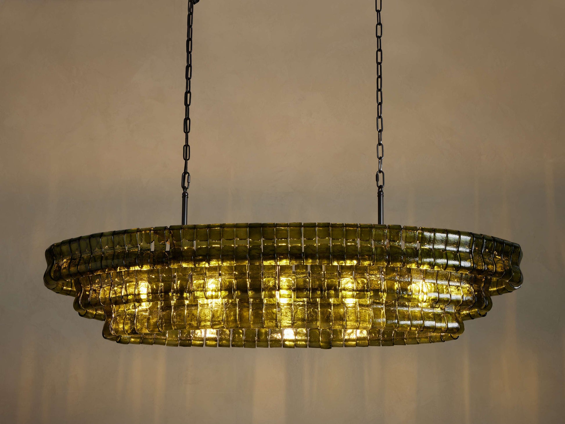 Ghiaccio Oval Chandelier, olive green curved glass squares in a tiered oval design, illuminated brightly, hanging from dark chains.